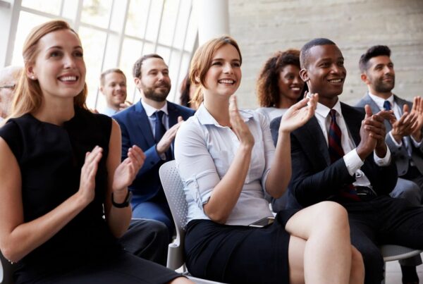 A group of people in a room all facing one direction clapping and smiling